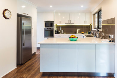 Exempel på ett mellanstort modernt vit vitt kök, med en nedsänkt diskho, släta luckor, vita skåp, brunt stänkskydd, rostfria vitvaror, en köksö, bänkskiva i kvarts, glaspanel som stänkskydd, laminatgolv och brunt golv