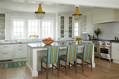 Example of a large beach style l-shaped medium tone wood floor eat-in kitchen design in New York with a farmhouse sink, recessed-panel cabinets, white cabinets, quartzite countertops, white backsplash, porcelain backsplash, stainless steel appliances and an island
