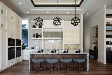 Example of a mid-sized transitional l-shaped dark wood floor and brown floor open concept kitchen design in Miami with a farmhouse sink, shaker cabinets, white cabinets, quartz countertops, beige backsplash, ceramic backsplash, black appliances, an island and gray countertops
