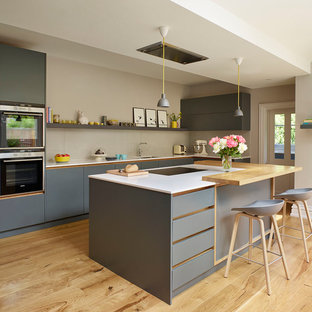 Photo of a large scandi l-shaped kitchen in London with flat-panel cabinets, grey cabinets, composite countertops, beige splashback, light hardwood flooring, an island, beige floors, white worktops, a submerged sink, glass sheet splashback and stainless steel appliances.