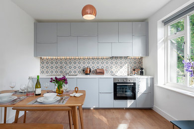 Small contemporary single-wall kitchen/diner in Wiltshire with flat-panel cabinets, blue cabinets, stainless steel appliances, light hardwood flooring, brown floors, a built-in sink and multi-coloured splashback.