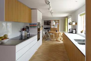 This is an example of a medium sized contemporary galley open plan kitchen in Hampshire with a submerged sink, flat-panel cabinets, light wood cabinets, stainless steel worktops, white splashback, glass sheet splashback, stainless steel appliances, light hardwood flooring, no island, beige floors and white worktops.