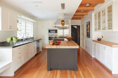 Inredning av ett klassiskt kök, med en integrerad diskho, skåp i shakerstil, vita skåp, bänkskiva i rostfritt stål, fönster som stänkskydd, rostfria vitvaror, mellanmörkt trägolv och en köksö