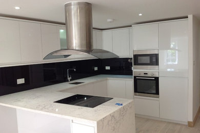 Medium sized contemporary u-shaped kitchen/diner in London with a built-in sink, glass-front cabinets, white cabinets, marble worktops, black splashback, glass sheet splashback, stainless steel appliances, light hardwood flooring and a breakfast bar.