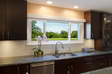 This is an example of a contemporary kitchen in Detroit with a double-bowl sink, flat-panel cabinets, dark wood cabinets, granite worktops, grey splashback, glass tiled splashback and stainless steel appliances.