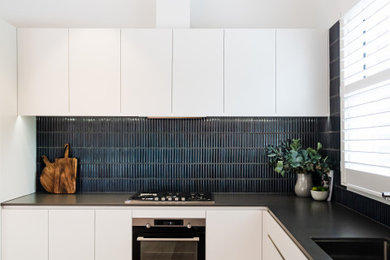 Inredning av ett modernt litet grå grått kök, med en undermonterad diskho, släta luckor, vita skåp, bänkskiva i kvarts, blått stänkskydd, stänkskydd i mosaik, svarta vitvaror och ljust trägolv