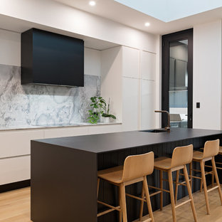 Photo of a contemporary galley kitchen in Adelaide with an integrated sink, flat-panel cabinets, white cabinets, grey splashback, black appliances, light hardwood floors, with island and black benchtop.