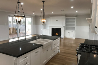 Inredning av ett lantligt svart svart kök, med en rustik diskho, skåp i shakerstil, vita skåp, granitbänkskiva, vitt stänkskydd, stänkskydd i keramik, svarta vitvaror, ljust trägolv, en köksö och brunt golv