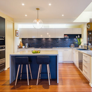 Large contemporary u-shaped open plan kitchen in Melbourne with flat-panel cabinets, quartz benchtops, black splashback, subway tile splashback, medium hardwood floors, with island, grey benchtop, a drop-in sink, black cabinets, black appliances and brown floor.