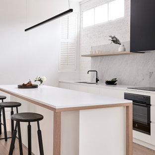 Inspiration for a contemporary galley kitchen in Melbourne with an undermount sink, flat-panel cabinets, white cabinets, grey splashback, stone slab splashback, black appliances, light hardwood floors, with island, beige floor and white benchtop.