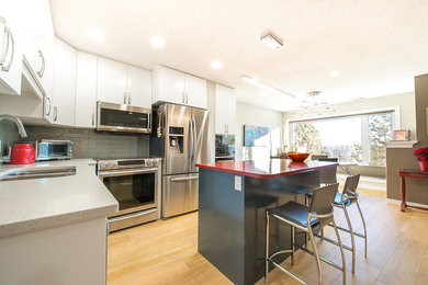 Eat-in kitchen - small contemporary l-shaped light wood floor and beige floor eat-in kitchen idea in Vancouver with an undermount sink, flat-panel cabinets, white cabinets, quartz countertops, gray backsplash, glass tile backsplash, stainless steel appliances and an island
