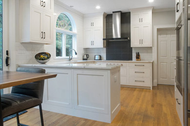 Mid-sized transitional u-shaped light wood floor and brown floor open concept kitchen photo in New York with an undermount sink, recessed-panel cabinets, white cabinets, quartz countertops, white backsplash, glass tile backsplash, stainless steel appliances, a peninsula and white countertops