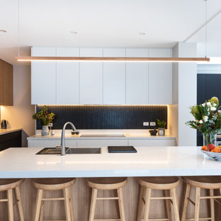 This is an example of a large contemporary kitchen in Sydney with a drop-in sink, flat-panel cabinets, quartz benchtops, black splashback, mosaic tile splashback, black appliances, light hardwood floors, with island and white benchtop.
