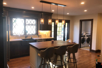 Inspiration for a mid-sized timeless l-shaped medium tone wood floor and brown floor eat-in kitchen remodel in New York with a farmhouse sink, raised-panel cabinets, dark wood cabinets, quartz countertops, beige backsplash, stone tile backsplash, stainless steel appliances, an island and white countertops