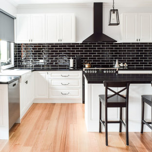 Photo of a large transitional l-shaped open plan kitchen in Melbourne with a farmhouse sink, white cabinets, quartz benchtops, black splashback, subway tile splashback, black appliances, with island, brown floor, raised-panel cabinets and medium hardwood floors.