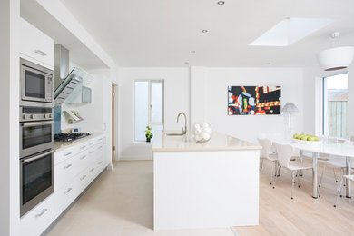 This is an example of a large contemporary u-shaped kitchen/diner in Dublin with a submerged sink, flat-panel cabinets, white cabinets, white splashback, stainless steel appliances, light hardwood flooring and an island.