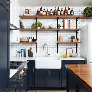 Small eclectic l-shaped open plan kitchen in DC Metro with a farmhouse sink, shaker cabinets, blue cabinets, quartz benchtops, white splashback, ceramic splashback, white appliances, porcelain floors, with island and brown floor.