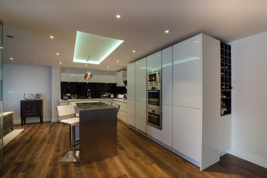 This is an example of a large contemporary l-shaped open plan kitchen in London with a built-in sink, flat-panel cabinets, white cabinets, black splashback, glass sheet splashback, stainless steel appliances, dark hardwood flooring and multiple islands.