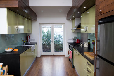 Cette image montre une petite cuisine américaine parallèle bohème avec un évier de ferme, un placard à porte plane, des portes de placards vertess et une crédence en feuille de verre.