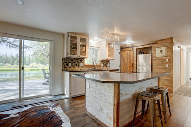 Example of a mid-sized mountain style l-shaped laminate floor and brown floor open concept kitchen design in Other with an undermount sink, glass-front cabinets, distressed cabinets, granite countertops, multicolored backsplash, ceramic backsplash, stainless steel appliances, an island and black countertops