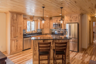 Example of a mid-sized mountain style l-shaped light wood floor open concept kitchen design in Minneapolis with a double-bowl sink, raised-panel cabinets, light wood cabinets, granite countertops, multicolored backsplash, matchstick tile backsplash, stainless steel appliances and an island