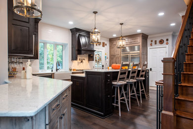 This is an example of a large rustic u-shaped kitchen/diner in Philadelphia with a belfast sink, raised-panel cabinets, medium wood cabinets, engineered stone countertops, white splashback, metro tiled splashback, stainless steel appliances, dark hardwood flooring, an island, brown floors and white worktops.