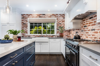 Example of a mid-sized country l-shaped dark wood floor and brown floor open concept kitchen design in Los Angeles with a farmhouse sink, shaker cabinets, white cabinets, quartz countertops, multicolored backsplash, brick backsplash, black appliances, an island and gray countertops