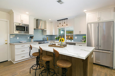 This is an example of a medium sized farmhouse l-shaped kitchen/diner in San Diego with a belfast sink, recessed-panel cabinets, white cabinets, blue splashback, metro tiled splashback, stainless steel appliances, porcelain flooring, an island, brown floors, white worktops, engineered stone countertops and exposed beams.