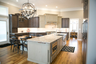 Large transitional u-shaped medium tone wood floor and brown floor kitchen photo in Indianapolis with a farmhouse sink, raised-panel cabinets, dark wood cabinets, an island, granite countertops, white backsplash, subway tile backsplash, paneled appliances and multicolored countertops