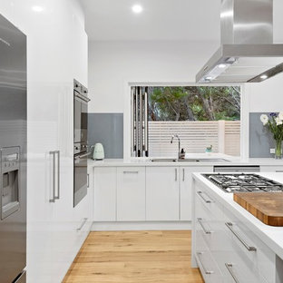 Design ideas for a beach style l-shaped kitchen in Melbourne with a drop-in sink, flat-panel cabinets, white cabinets, grey splashback, glass sheet splashback, stainless steel appliances, light hardwood floors, with island, beige floor and white benchtop.