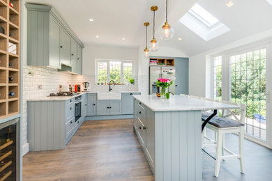 Design ideas for a medium sized farmhouse l-shaped kitchen/diner in Hertfordshire with a belfast sink, shaker cabinets, grey cabinets, marble worktops, white splashback, stainless steel appliances, medium hardwood flooring, an island, brown floors and metro tiled splashback.