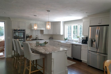 Example of a large transitional l-shaped medium tone wood floor and brown floor open concept kitchen design in Boston with a farmhouse sink, shaker cabinets, white cabinets, granite countertops, white backsplash, stainless steel appliances and an island
