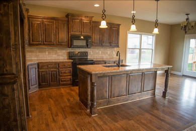 Example of a mid-sized classic l-shaped medium tone wood floor eat-in kitchen design in Kansas City with an undermount sink, distressed cabinets, granite countertops, beige backsplash, stone tile backsplash, black appliances, an island and raised-panel cabinets