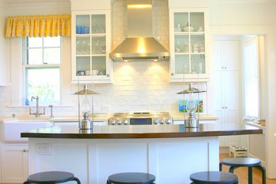 Inspiration for a small coastal l-shaped light wood floor enclosed kitchen remodel in Boston with a farmhouse sink, glass-front cabinets, white cabinets, wood countertops, white backsplash, stone tile backsplash, stainless steel appliances and an island