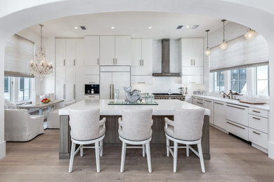 Example of a mid-sized transitional l-shaped light wood floor open concept kitchen design in Orange County with flat-panel cabinets, white cabinets, solid surface countertops, metallic backsplash, an island, an undermount sink, glass tile backsplash and white appliances