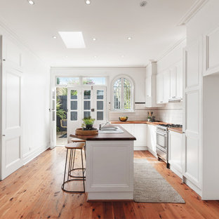 This is an example of a country u-shaped kitchen in Melbourne with a drop-in sink, recessed-panel cabinets, white cabinets, wood benchtops, white splashback, subway tile splashback, stainless steel appliances, medium hardwood floors, with island, brown floor and brown benchtop.