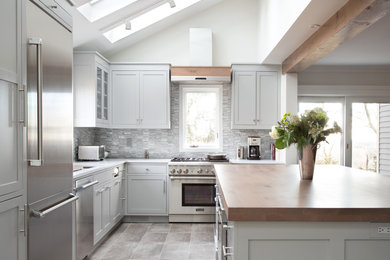 Inspiration for a mid-sized transitional l-shaped porcelain tile open concept kitchen remodel in New York with an undermount sink, shaker cabinets, gray cabinets, wood countertops, gray backsplash, stone tile backsplash, stainless steel appliances and an island