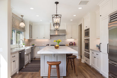 Mid-sized transitional u-shaped dark wood floor and brown floor kitchen photo in Los Angeles with an island, a farmhouse sink, recessed-panel cabinets, white cabinets, marble countertops, stainless steel appliances, white countertops, gray backsplash and subway tile backsplash