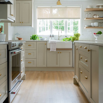 Example of a mid-sized transitional l-shaped light wood floor and brown floor kitchen design in Boston with a farmhouse sink, shaker cabinets, beige cabinets, marble countertops, white backsplash, subway tile backsplash, stainless steel appliances, an island and white countertops