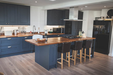 Photo of a medium sized rustic single-wall open plan kitchen in Devon with a belfast sink, shaker cabinets, grey cabinets, wood worktops, black appliances, laminate floors and a breakfast bar.