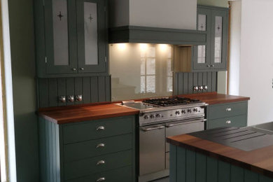 This is an example of a country open plan kitchen with shaker cabinets, green cabinets, wood worktops, green splashback, stainless steel appliances, an island and brown worktops.