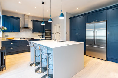 Design ideas for a medium sized contemporary l-shaped open plan kitchen in London with an integrated sink, shaker cabinets, blue cabinets, composite countertops, stainless steel appliances, light hardwood flooring, an island, beige floors and white worktops.