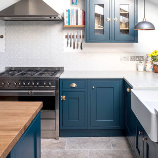 Medium sized farmhouse u-shaped open plan kitchen in London with a belfast sink, shaker cabinets, blue cabinets, white splashback, porcelain splashback, stainless steel appliances, porcelain flooring, an island, white worktops, composite countertops and grey floors.