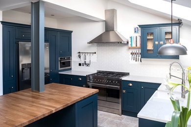 Design ideas for a medium sized farmhouse u-shaped open plan kitchen in Surrey with a belfast sink, shaker cabinets, blue cabinets, white splashback, porcelain splashback, stainless steel appliances, travertine flooring, an island, white worktops, composite countertops and grey floors.