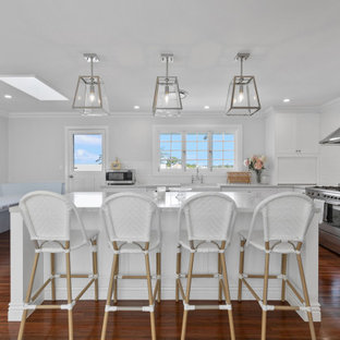 Photo of a beach style kitchen in Brisbane with a farmhouse sink, shaker cabinets, white cabinets, white splashback, stainless steel appliances, medium hardwood floors, with island and grey benchtop.
