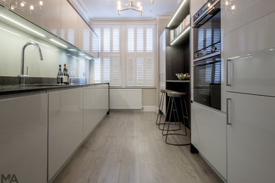 Design ideas for a large contemporary galley open plan kitchen in London with a submerged sink, flat-panel cabinets, grey cabinets, quartz worktops, grey splashback, glass sheet splashback, black appliances, light hardwood flooring and grey floors.