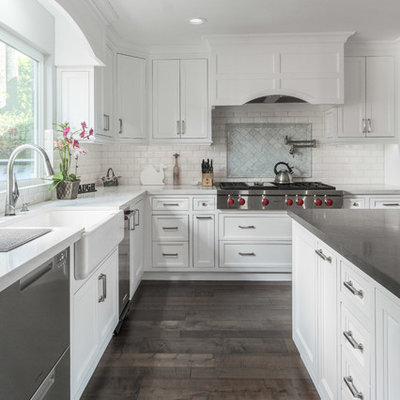 Transitional l-shaped dark wood floor and brown floor kitchen photo in Los Angeles with a farmhouse sink, recessed-panel cabinets, white cabinets, white backsplash, subway tile backsplash, stainless steel appliances, an island and white countertops