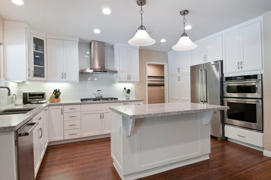 This is an example of a medium sized traditional u-shaped kitchen in Los Angeles with a double-bowl sink, shaker cabinets, white cabinets, granite worktops, white splashback, glass tiled splashback, stainless steel appliances, medium hardwood flooring, an island and brown floors.