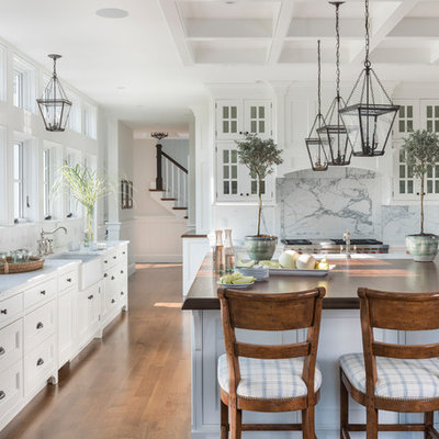 Example of a beach style medium tone wood floor kitchen design in Boston with a farmhouse sink, white cabinets, white backsplash, stone slab backsplash, stainless steel appliances, an island, white countertops and recessed-panel cabinets