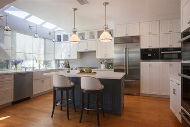 Example of a mid-sized trendy l-shaped light wood floor and brown floor open concept kitchen design in Los Angeles with a farmhouse sink, shaker cabinets, white cabinets, marble countertops, gray backsplash, subway tile backsplash, stainless steel appliances and an island